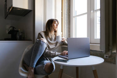 A Woman Enjoys A Calm Moment With A Cup Of Coffee As She Works On Her Laptop In A Cozy Home Setting, Bathed In Natural Light By The Window